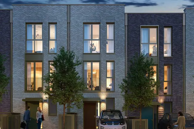 Kidbrooke Village’daki The Blackheath Collection’da sokak ağaçları ve park edilmiş arabalarla üç katlı sıra evlerin akşam görünümü.
