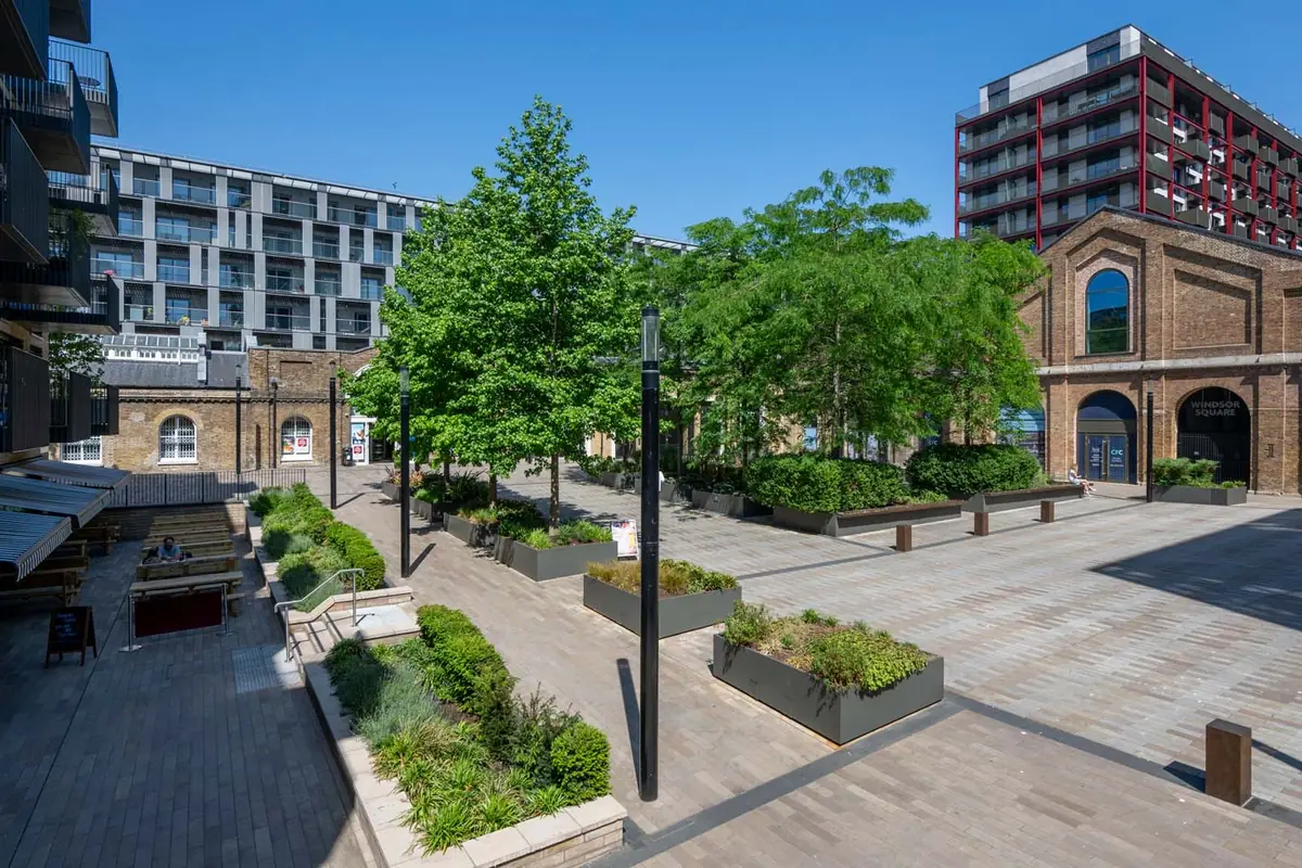 Royal Arsenal Riverside’da apartman binaları ile tarihi tuğla binalar arasında ağaçlı ve saksılı, taş döşeli bir avlu.