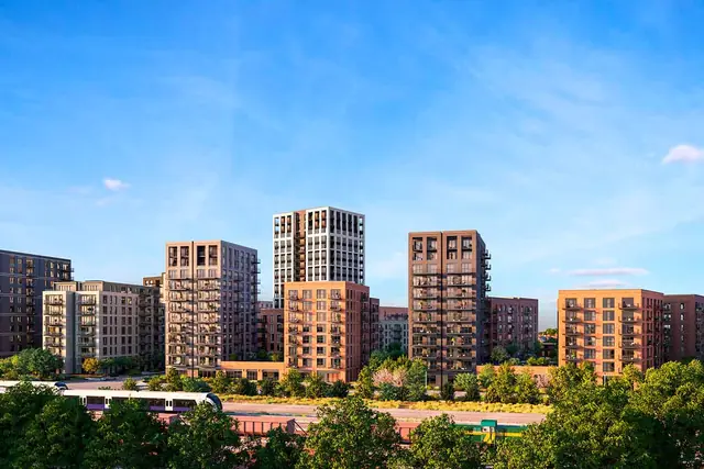 A row of brick apartment buildings at The Green Quarter behind trees and a railway line, under a blue sky.