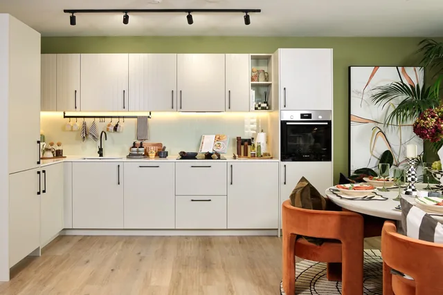 A white fitted kitchen with a dining table and orange chairs in the two-bedroom show apartment at The Green Quarter.