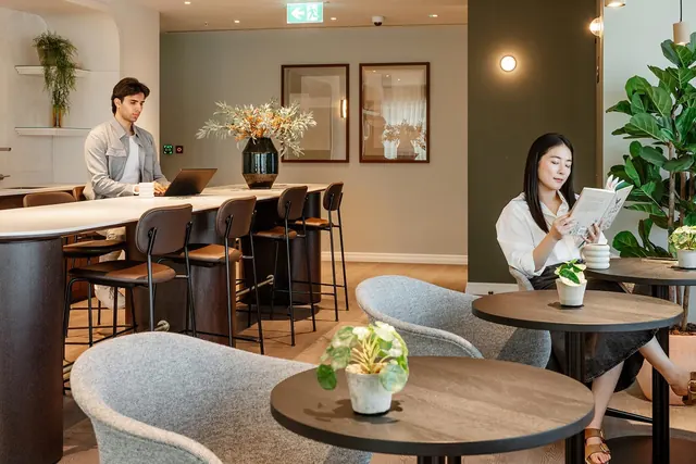 Two people working and reading in a lounge with a long counter, high stools and round tables at TwelveTrees Park.