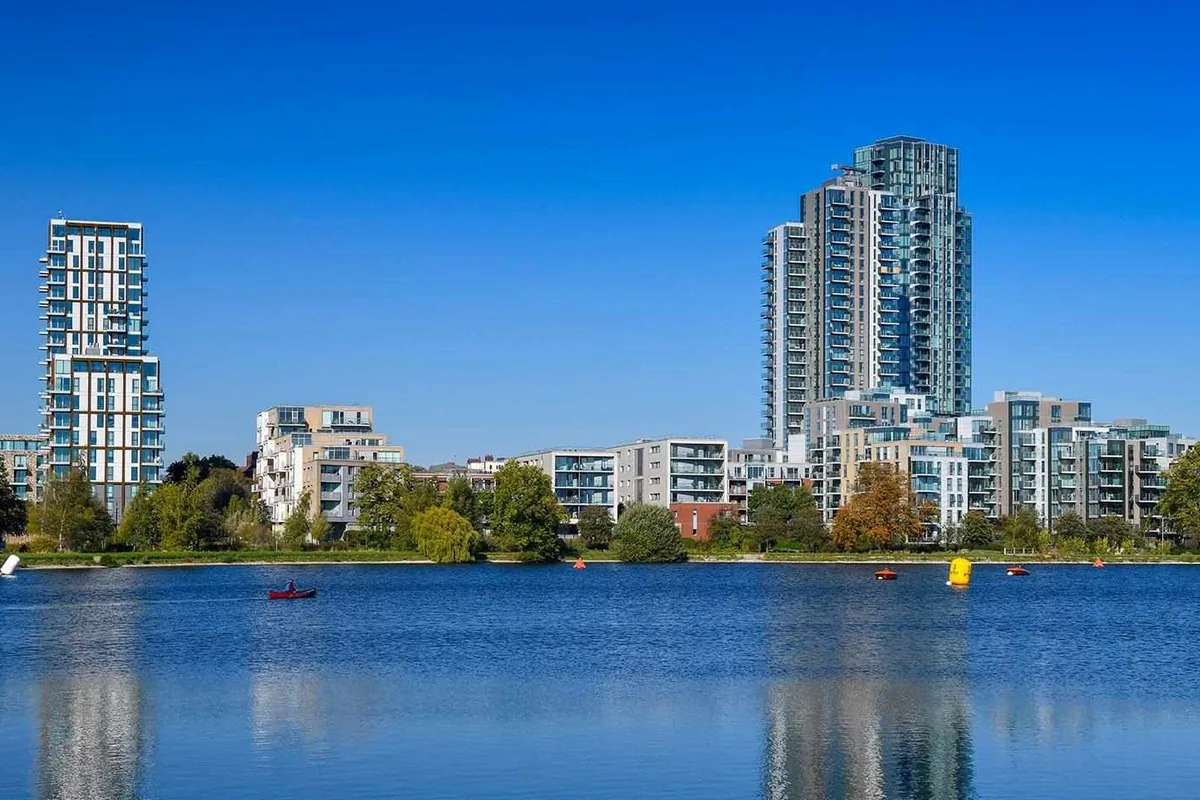 Apartment buildings and a tall tower at Woodberry Down seen across the water under a clear blue sky.