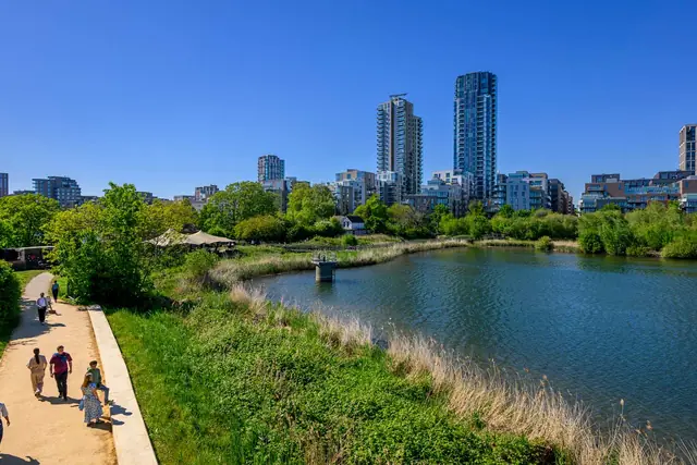 People walking on a path beside the water at Woodberry Down, with reeds, trees and residential towers behind.