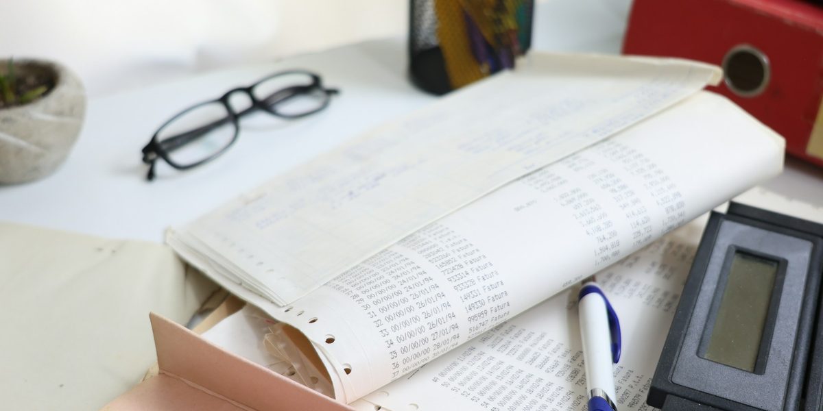 A desk with printed papers, reading glasses, a pen and a calculator